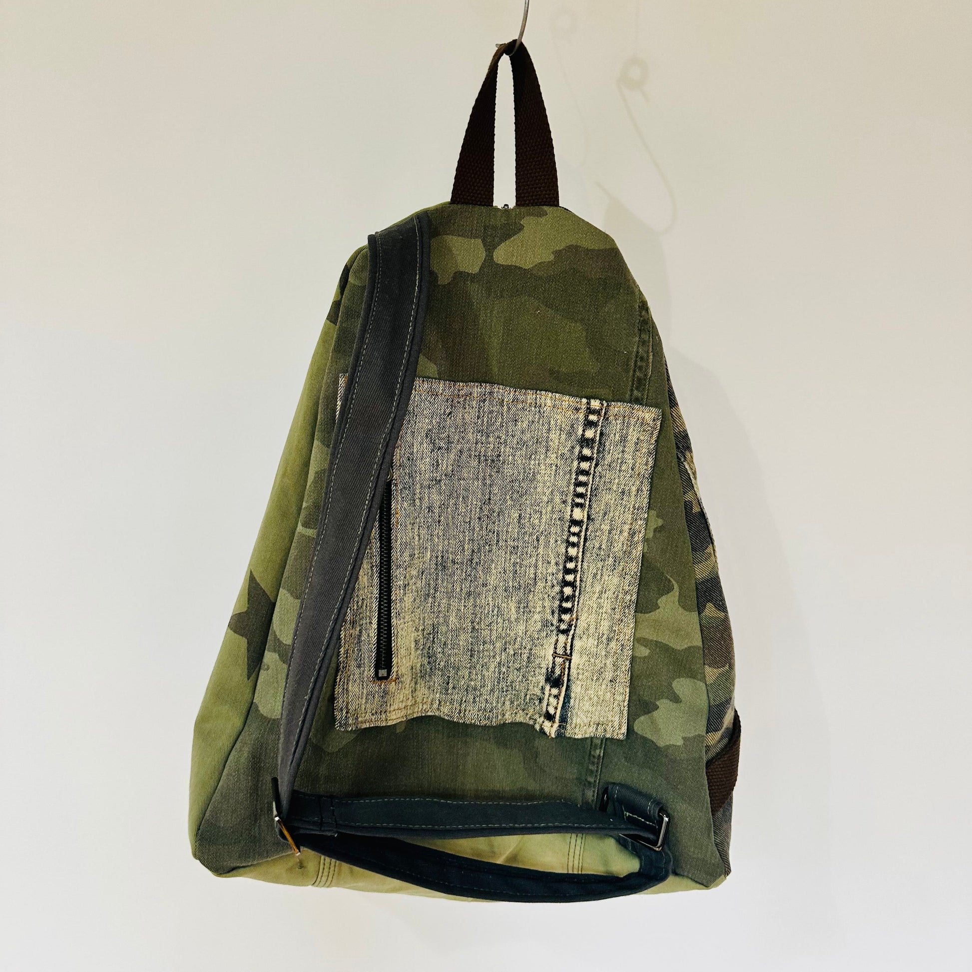 A canvas backpack with a green camouflage pattern, featuring a large front patch pocket with a woven beige fabric insert and dark straps, hanging against a plain white wall.