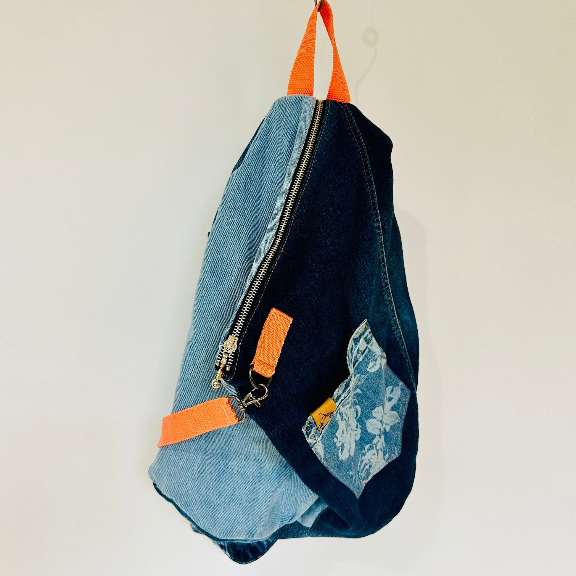 A handmade denim backpack hanging from a metal hook against a plain off-white wall. The bag features a mix of light and dark blue denim panels, a floral patterned pocket, and bright orange webbed accents on the top handles and zipper pulls.